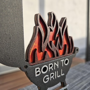 K&ouml;nnte beinhalten: Ein Holzschild mit schwarzem Hintergrund und dem Text "Born to Grill" in Wei&szlig;. Das Schild hat ein mehrschichtiges Holzflammen-Design mit roten und orangen Akzenten.