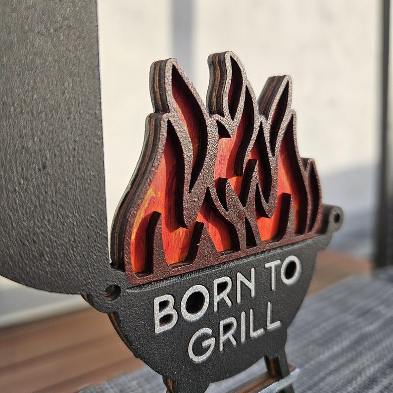 K&ouml;nnte beinhalten: Ein Holzschild mit schwarzem Hintergrund und dem Text "Born to Grill" in Wei&szlig;. Das Schild hat ein mehrschichtiges Holzflammen-Design mit roten und orangen Akzenten.