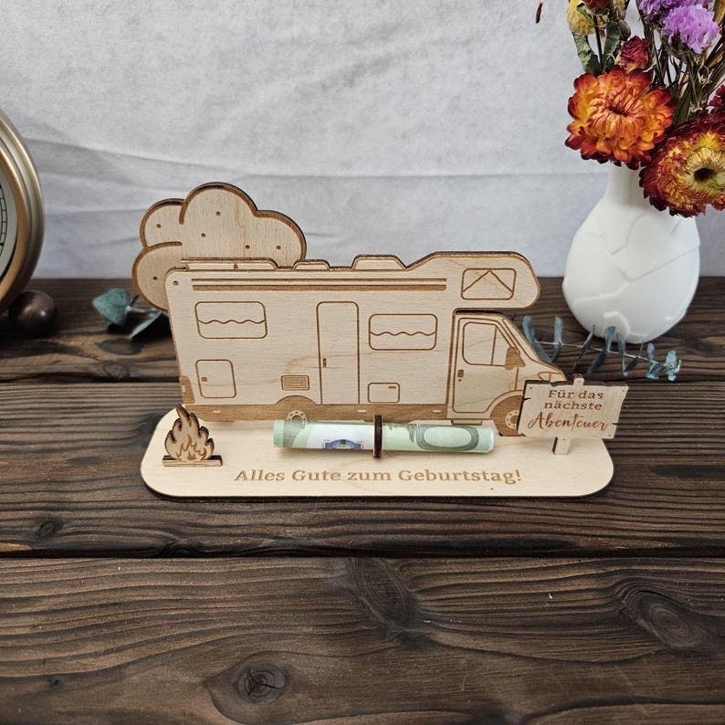 May include: A wooden campervan-shaped gift holder with a rolled-up banknote in front. The holder features a cloud and flame design, with German text reading "Alles Gute zum Geburtstag!" and "F&uuml;r das n&auml;chste Abenteuer."