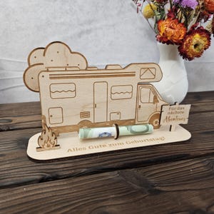 May include: A wooden campervan-shaped gift holder with a rolled-up banknote secured by a black band. The holder has a cloud and flame design, with the text "Alles Gute zum Geburtstag!" and "F&uuml;r das n&auml;chste Abenteuer".