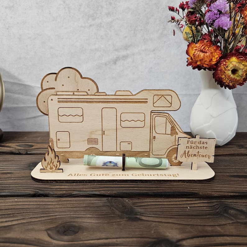 May include: A wooden campervan-shaped money holder with a cloud and flame design. The holder has the German text "Alles Gute zum Geburtstag!" and "F&uuml;r das n&auml;chste Abenteuer". A rolled-up banknote is held in place.