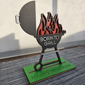 K&ouml;nnte beinhalten: Ein Grill aus Holz mit Flammen und dem Text "Born to Grill" auf der Vorderseite. Der Grill steht auf einem gr&uuml;nen Sockel mit dem Text "Happy Birthday".