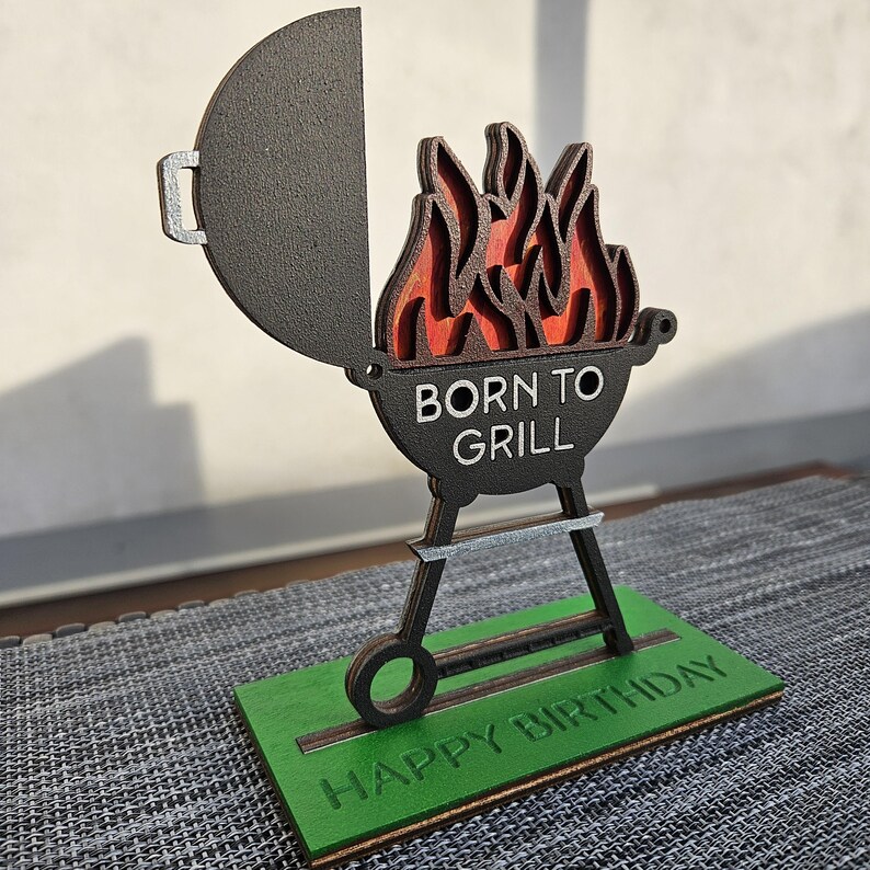 K&ouml;nnte beinhalten: Ein Grill aus Holz mit Flammen und dem Text "Born to Grill" auf der Vorderseite. Der Grill steht auf einem gr&uuml;nen Sockel mit dem Text "Happy Birthday".