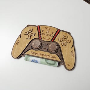Könnte beinhalten: Ein Geschenkhalter in Form eines Holz-Gamecontrollers mit dem Text "LEVEL 18 UNLOCKED" und "Happy Birthday Lucas". Der Halter besteht aus hellem und dunklem braunem Holz und hält einen aufgerollten Geldschein.