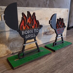 K&ouml;nnte beinhalten: Zwei h&ouml;lzerne Grill-f&ouml;rmige Geburtstagsdekorationen mit dem Text "Born to Grill" und einem Flammen-Design. Die Dekorationen stehen auf einem gr&uuml;nen Sockel mit dem Text "Happy Birthday Michael" und "Happy Birthday in June".