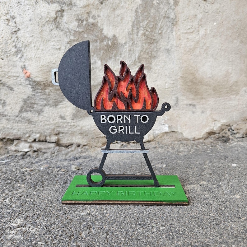 K&ouml;nnte beinhalten: Ein schwarzer und roter Grill aus Holz mit Flammen und dem Text "Born to Grill" auf der Vorderseite. Der Grill steht auf einem gr&uuml;nen Sockel mit dem Text "Happy Birthday".