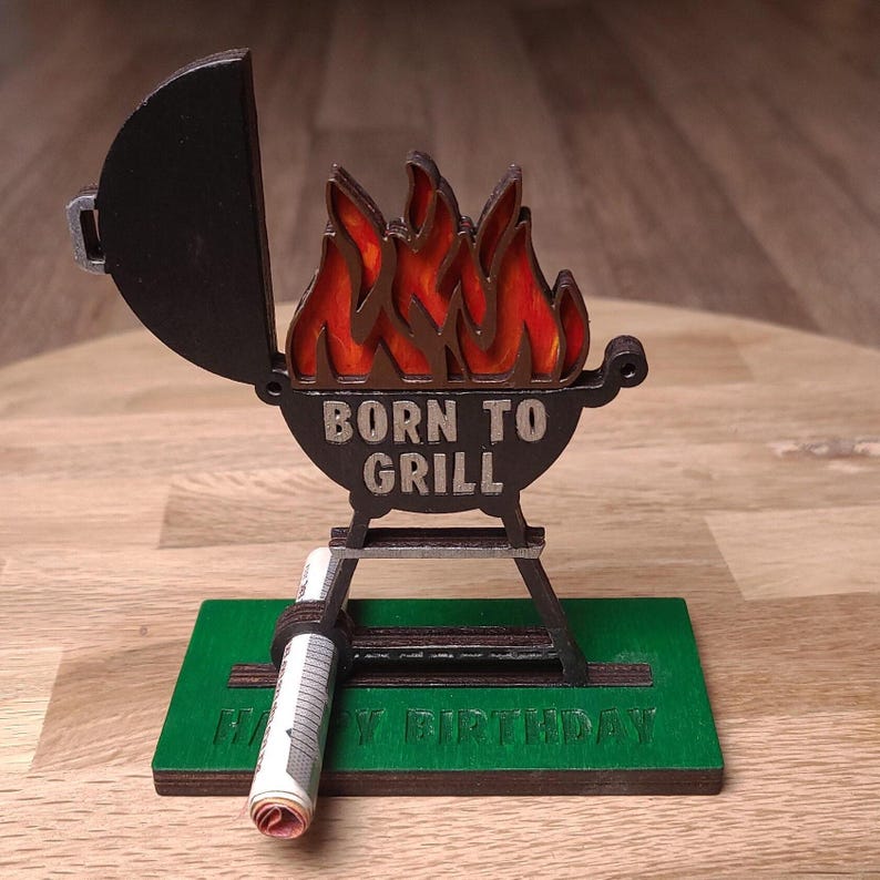 K&ouml;nnte beinhalten: Ein Grill aus Holz mit einem roten und orangenen Flammenmuster und dem Text "Born to Grill". Der Grill steht auf einem St&auml;nder mit einer gr&uuml;nen Basis, auf der "Happy Birthday" steht.