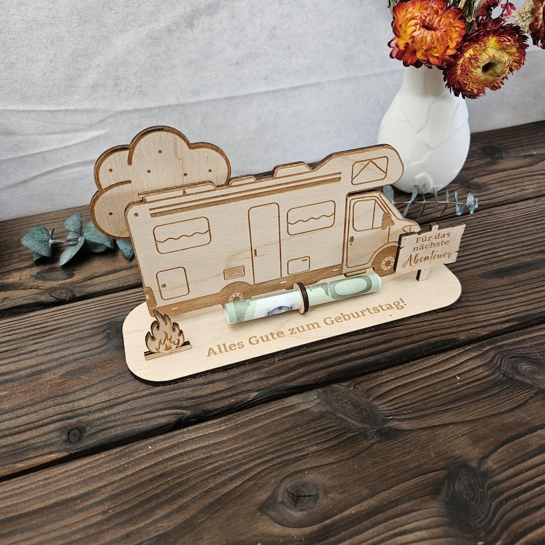 May include: A wooden campervan-shaped money holder with a cloud and flame design. The holder has the text "Alles Gute zum Geburtstag!" and "F&uuml;r das n&auml;chste Abenteuer" engraved. A rolled-up banknote is held in place by a brown band.
