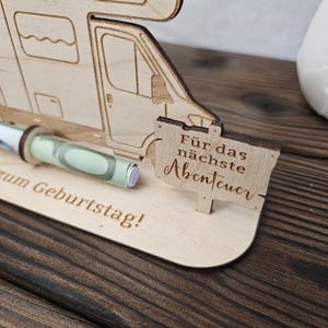 May include: A wooden money holder shaped like a camper van, with a rolled-up banknote visible. A small sign reads "F&uuml;r das n&auml;chste Abenteuer" (For the next adventure). The base has the text "zum Geburtstag!" (Happy Birthday!).