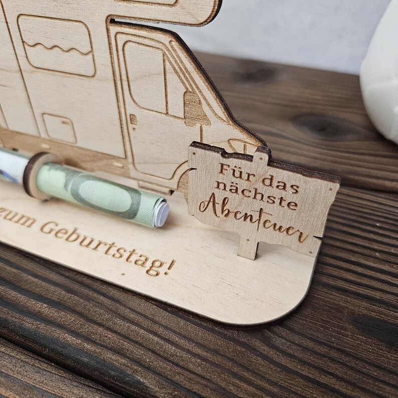 May include: A wooden money holder shaped like a camper van, with a rolled-up banknote visible. A small sign reads "F&uuml;r das n&auml;chste Abenteuer" (For the next adventure). The base has the text "zum Geburtstag!" (Happy Birthday!).