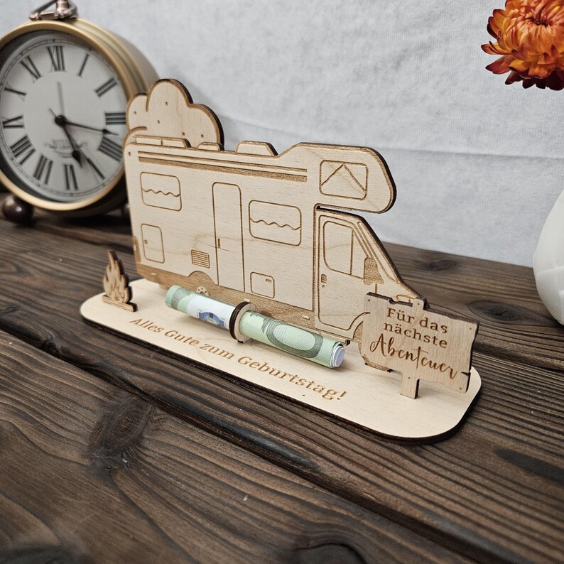 May include: A wooden campervan-shaped money holder with the text "Alles Gute zum Geburtstag!" and "F&uuml;r das n&auml;chste Abenteuer". The holder has a slot for rolled-up banknotes. A vintage clock and orange flower are in the background.