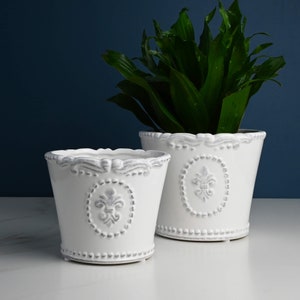 May include: Two white ceramic flower pots with a decorative grey fleur-de-lis design. The larger pot is taller and wider than the smaller pot.
