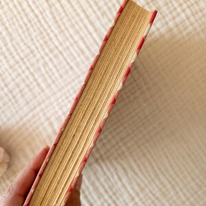 May include: A vintage book with aged, yellowed pages. The book's spine and edges are adorned with a red and white patterned fabric. The book is held in a hand, showcasing its well-worn condition and classic design.