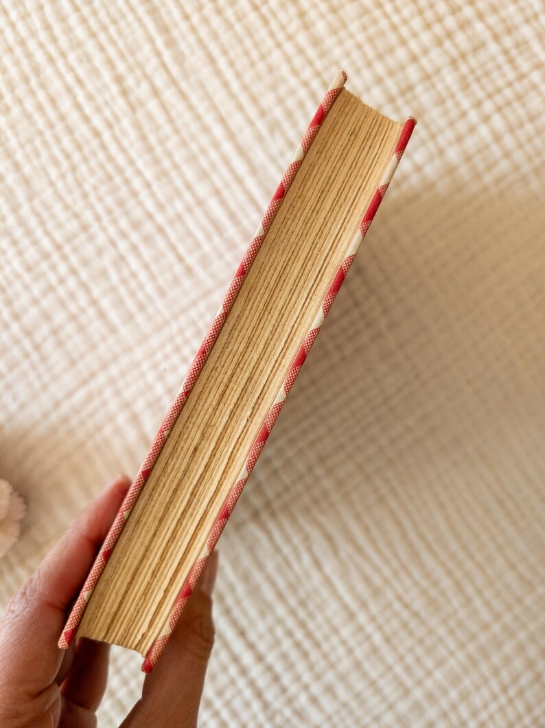 May include: A vintage book with aged, yellowed pages. The book's spine and edges are adorned with a red and white patterned fabric. The book is held in a hand, showcasing its well-worn condition and classic design.