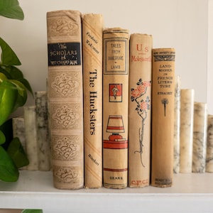 May include: A collection of vintage books with aged covers, displayed on a shelf. Titles include "The Scholars", "The Hucksters", "Tales From Shakespeare", and "Landmarks in French Literature". The books are in varying shades of beige and tan.