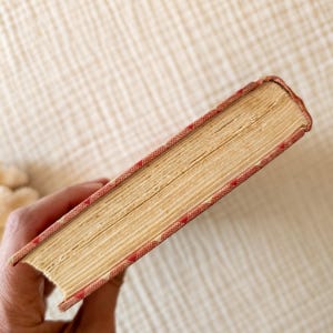 May include: A vintage book with aged, yellowed pages. The cover edges are red with a repeating white diamond pattern. The book's spine is visible, and the pages are slightly worn, suggesting age and use. The book is held in a hand.