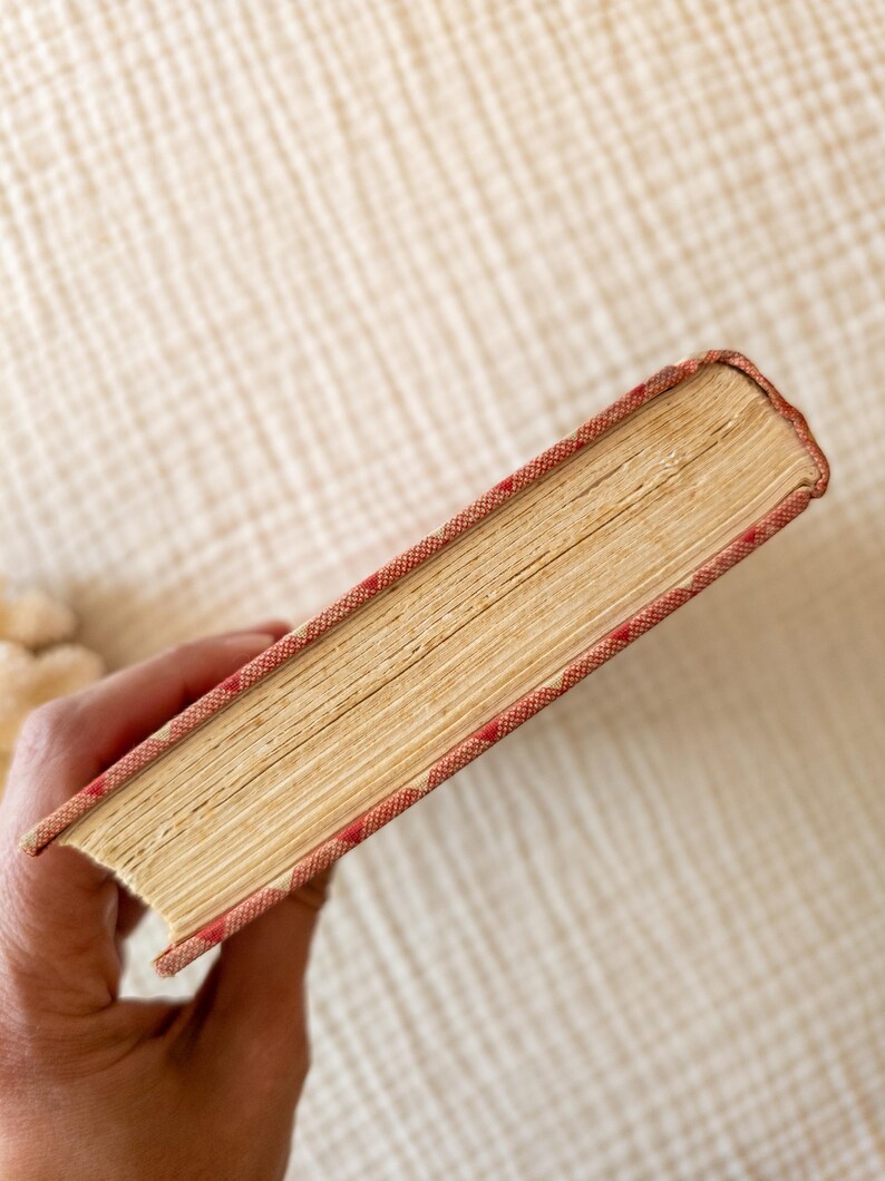 May include: A vintage book with aged, yellowed pages. The cover edges are red with a repeating white diamond pattern. The book's spine is visible, and the pages are slightly worn, suggesting age and use. The book is held in a hand.