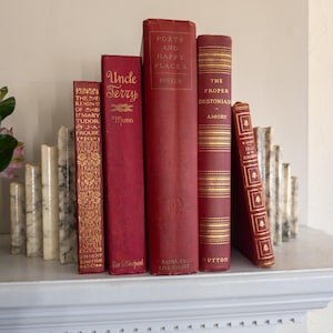 May include: A collection of five vintage hardcover books with red spines and gold lettering, arranged between two white marble bookends. The titles include The Reign of Mary Tudor, Uncle Terry, Ports and Happy Places, The Proper Bostonian, and Tales.