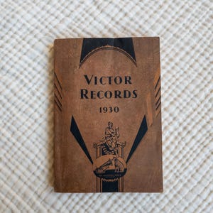 May include: A brown book with a black and white design. The book is titled "Victor Records" and the year "1930" is printed on the cover. The book features a black and white illustration of a person holding a microphone and a dog.