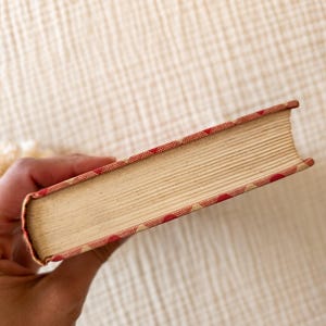 May include: A vintage book with aged, cream-colored pages. The book's edges are adorned with a red and white patterned design. The book is held in a hand, showcasing the side profile of the book.