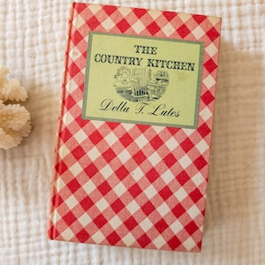 May include: A vintage cookbook titled "The Country Kitchen" by Della T. Lutes. The book has a red and white checkered cover and a green rectangular label with the title and author's name. The book is on a white, textured surface.