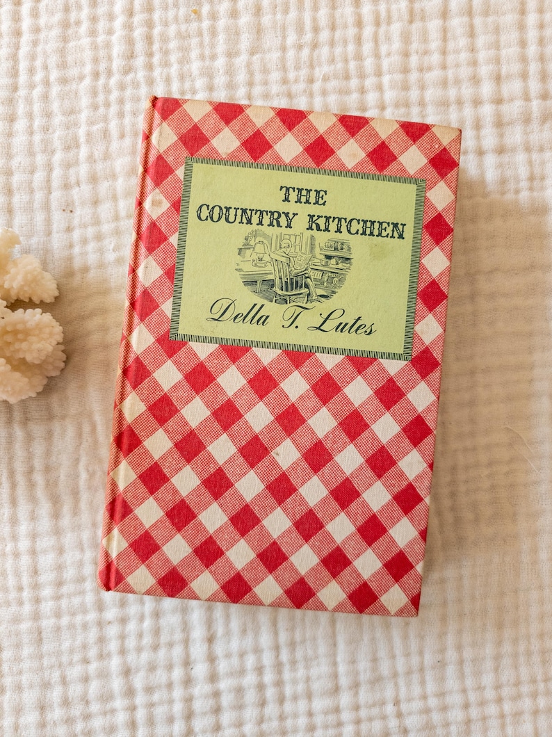 May include: A vintage cookbook titled "The Country Kitchen" by Della T. Lutes. The book has a red and white checkered cover and a green rectangular label with the title and author's name. The book is on a white, textured surface.