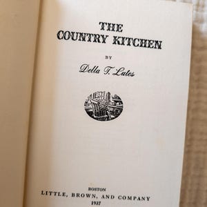 May include: Open book page with the title "THE COUNTRY KITCHEN" in bold black letters. Below the title is "BY" and the author's name, Della T. Lutes. A small circular illustration is centered below the author's name. The publisher and year, 1937, are at the bottom.