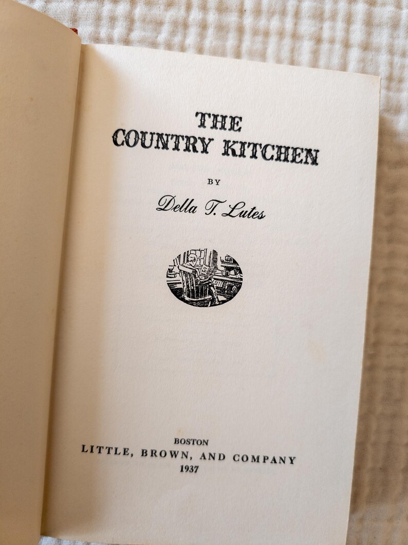 May include: Open book page with the title "THE COUNTRY KITCHEN" in bold black letters. Below the title is "BY" and the author's name, Della T. Lutes. A small circular illustration is centered below the author's name. The publisher and year, 1937, are at the bottom.