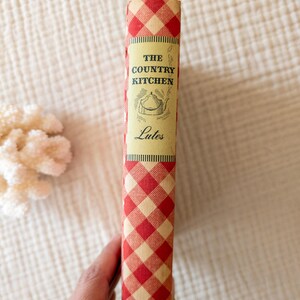 May include: A vintage cookbook titled "The Country Kitchen" with a red and cream checkered spine. The book's cover features an illustration of a teapot. The book is a vintage item, likely from the mid-20th century.