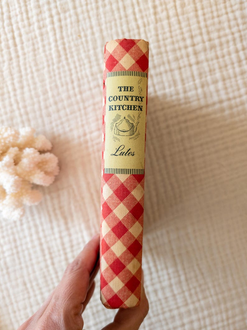 May include: A vintage cookbook titled "The Country Kitchen" with a red and cream checkered spine. The book's cover features an illustration of a teapot. The book is a vintage item, likely from the mid-20th century.