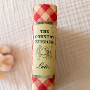 May include: A vintage cookbook titled "The Country Kitchen" with a red and white checkered pattern on the spine. The book's cover features a cream-colored label with the title, a teapot illustration, and the name "Lutes".