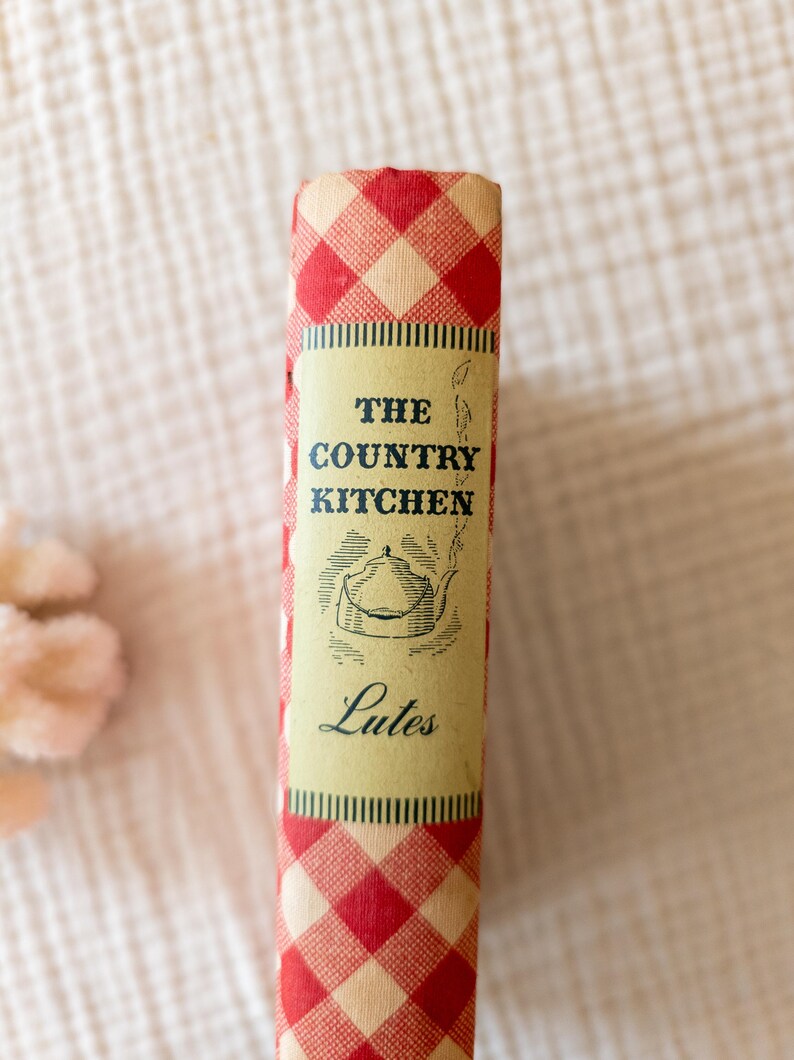 May include: A vintage cookbook titled "The Country Kitchen" with a red and white checkered pattern on the spine. The book's cover features a cream-colored label with the title, a teapot illustration, and the name "Lutes".