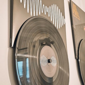 Puede incluir: Un disco de vinilo negro con un patrón de ondas blancas en la etiqueta está montado en una pared junto a otro disco con un gráfico en blanco y negro.
