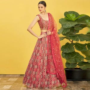 May include: A red and gold embroidered lehenga with a matching dupatta. The sleeveless top is heavily embellished with gold thread and sequins. The skirt features a geometric pattern in shades of red, gold, and brown. A sheer red dupatta is draped over the shoulder.