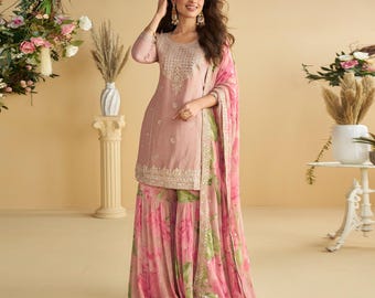 Op de afbeelding: Een blush roze Indiase salwar kameez set. De top heeft ingewikkelde borduursels en een ronde halslijn. De broek met wijde pijpen en de dupatta hebben een bloemenprint met roze en groene tinten. De outfit is geaccessoireerd met gouden sieraden.