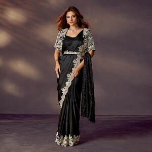 May include: A black satin saree with intricate floral embroidery along the border and a matching embellished bolero jacket. The outfit includes a decorative belt and a sequined shawl, creating an elegant ensemble.