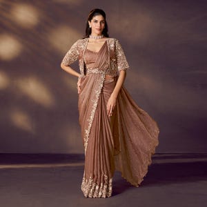 May include: A brown saree with intricate embroidery and a matching bolero jacket. The saree features delicate floral patterns along the border and scattered across the sheer fabric. The bolero is adorned with shimmering embellishments.