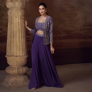 Pode incluir: Um conjunto roxo de três peças com um top curto, calças palazzo e um casaco. O top curto e o casaco são adornados com bordados intrincados. O casaco tem decote quadrado e mangas três quartos. As calças palazzo têm um design de perna larga.
