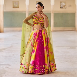 May include: A vibrant, multi-colored lehenga ensemble. The skirt features a tie-dye pattern in yellow, pink, and green. The cropped top is adorned with floral embellishments. A sheer, light green shawl completes the look. The outfit is suitable for festive occasions.
