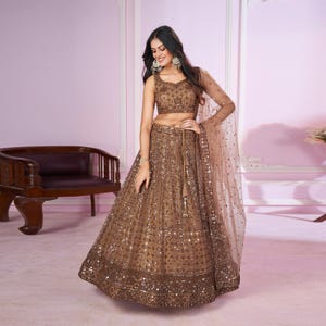 May include: A brown lehenga choli set with intricate embellishments. The set features a sleeveless crop top and a long, flared skirt, both decorated with detailed patterns. A sheer dupatta with matching embellishments completes the outfit. Suitable for special occasions.