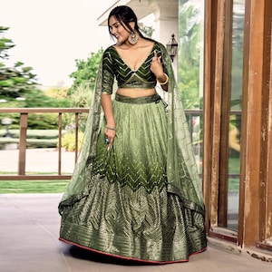 May include: A two-piece lehenga set in shades of olive and forest green. The cropped blouse features a deep V-neckline and short sleeves with sequin embroidery. The matching pleated skirt has a chevron pattern and a wide, heavily embellished hem.