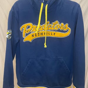 May include: A blue hoodie with a yellow and white striped band at the bottom. The hoodie has a yellow drawstring and a large yellow and white embroidered logo that says "Predators Nashville".
