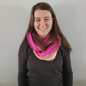 May include: A handmade crochet infinity scarf in shades of pink, magenta, and cream. The scarf is worn around the neck and has a striped pattern. The yarn appears to be soft and the scarf is likely warm. The scarf is a fashion accessory.