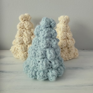 May include: Three handmade crochet Christmas trees in cream and light blue. The trees are made with a textured stitch, giving them a bubbly appearance. The trees are arranged on a white surface, with a light gray background.