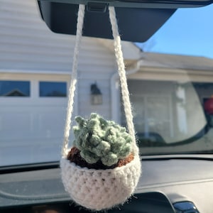 May include: A white crocheted hanging planter with a green crocheted succulent inside. The planter is hanging from a rearview mirror in a car.