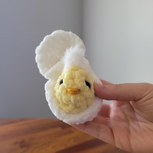 May include: A crocheted chick toy in a white eggshell. The chick is yellow with black eyes and an orange beak.