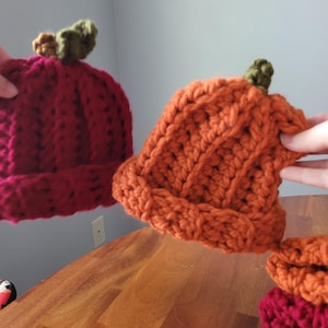 May include: Handmade crochet pumpkin hats in shades of orange and burgundy. Each hat features a green stem detail. The hats are made with a chunky yarn, perfect for fall and Halloween.