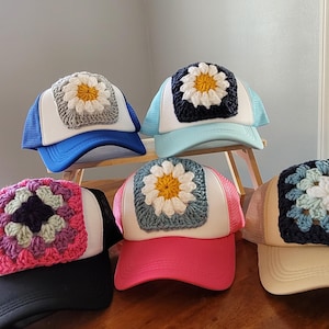 May include: Five trucker hats featuring white front panels and mesh backs in various colors. Each hat displays a square crochet granny square patch on the front, with four featuring a white and yellow daisy design and one featuring a multi-colored floral pattern.