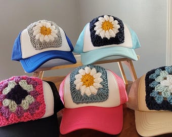 May include: Five trucker hats featuring white front panels and mesh backs in various colors. Each hat displays a square crochet granny square patch on the front, with four featuring a white and yellow daisy design and one featuring a multi-colored floral pattern.