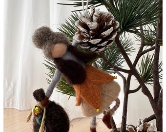 May include: A handmade felt Christmas ornament depicting a whimsical gnome figure. The gnome wears a gray hat, brown vest, and orange skirt, holding a sack with a golden bell. The ornament is suspended near a pine cone and evergreen branches.
